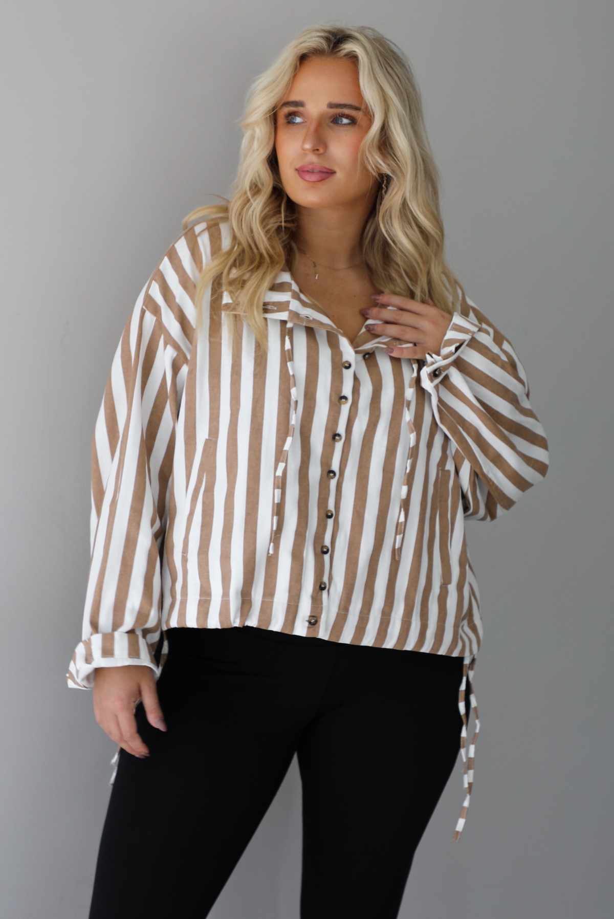 A woman wearing a tan and white striped collared long sleeved top with functional button closures along the front, worn with black leggings, standing indoors against a white wall. The tops drawstring details are un-cinched to show the full length. 