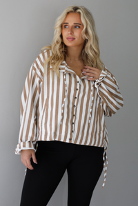 A woman wearing a tan and white striped collared long sleeved top with functional button closures along the front, worn with black leggings, standing indoors against a white wall. The tops drawstring details are un-cinched to show the full length. 