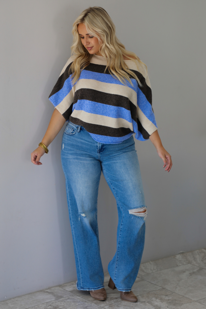 A woman wearing a cream, brown,, and blue striped knit top that has a boat-neck design, and quarter length flowy sleeves, paired with a pair of medium wash denim jeans, standing indoors against a white wall with white marble flooring. 