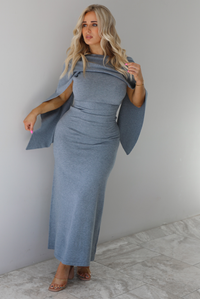 Woman wearing a grey maxi length knit dress that has a high neckline with an attached shawl detail that covers the upper half of the chest and flows on either shoulder in the back. The dress is fitted with a slightly flowy lower hem. The dress is paired with clear strapped nude heels. The photo is taken indoors against a white wall with white marble flooring. 