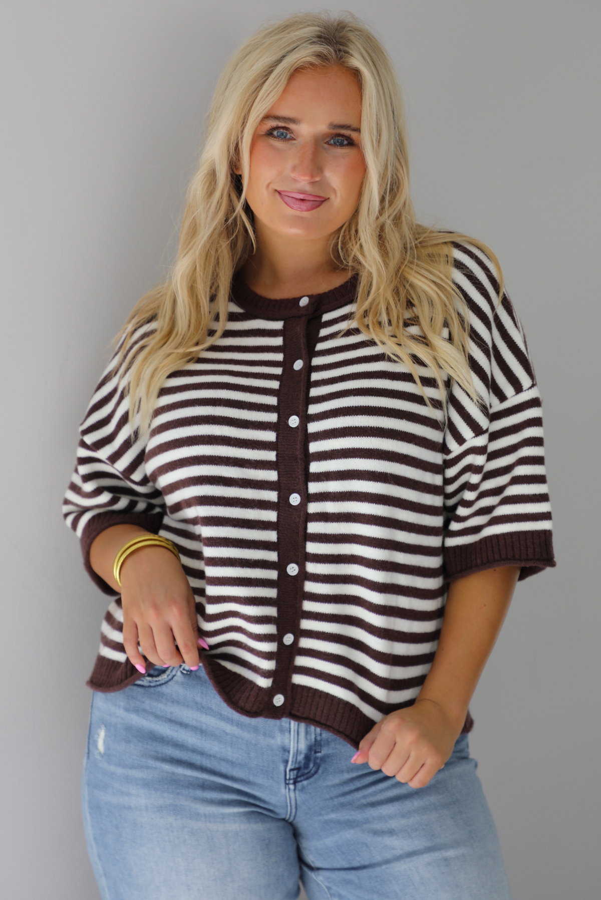 A woman wearing a brown and white striped knit button top cardigan top, paired with light wash denim distressed denim, standing indoors against a white wall.