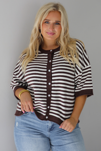 A woman wearing a brown and white striped knit button top cardigan top, paired with light wash denim distressed denim, standing indoors against a white wall.
