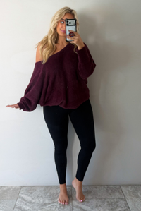 A woman taking a mirror selfie wearing a maroon oversized long sleeved sweater, worn off the shoulder, paired with black leggings, indoors against a white wall with white marble flooring. 