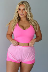 A woman modeling a bright pink two-piece activewear set consisting of a scoop-neck crop top and matching athletic shorts with a darker, hot pink trim, standing indoors against a white wall.