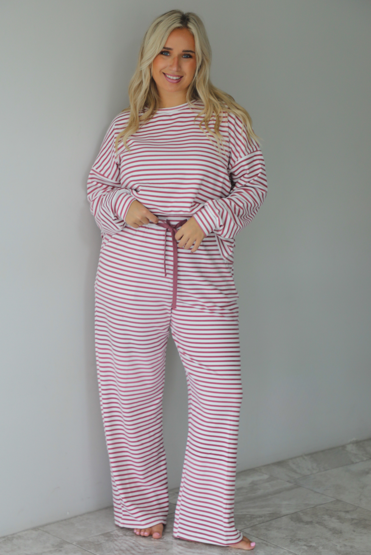A woman wearing a matching two piece set featuring a long sleeved top and high waisted wide leg pants with a berry and white striped pattern throughout, standing indoors against a white wall with white marble flooring. 