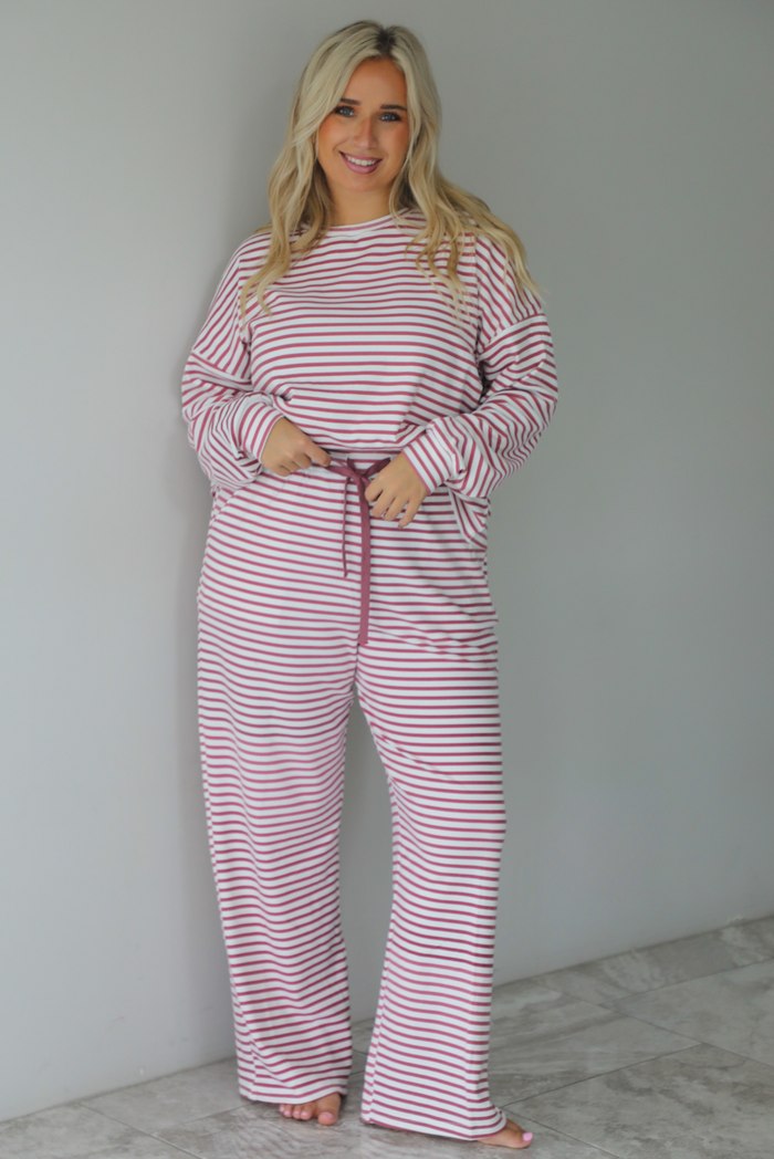 A woman wearing a matching two piece set featuring a long sleeved top and high waisted wide leg pants with a berry and white striped pattern throughout, standing indoors against a white wall with white marble flooring. 