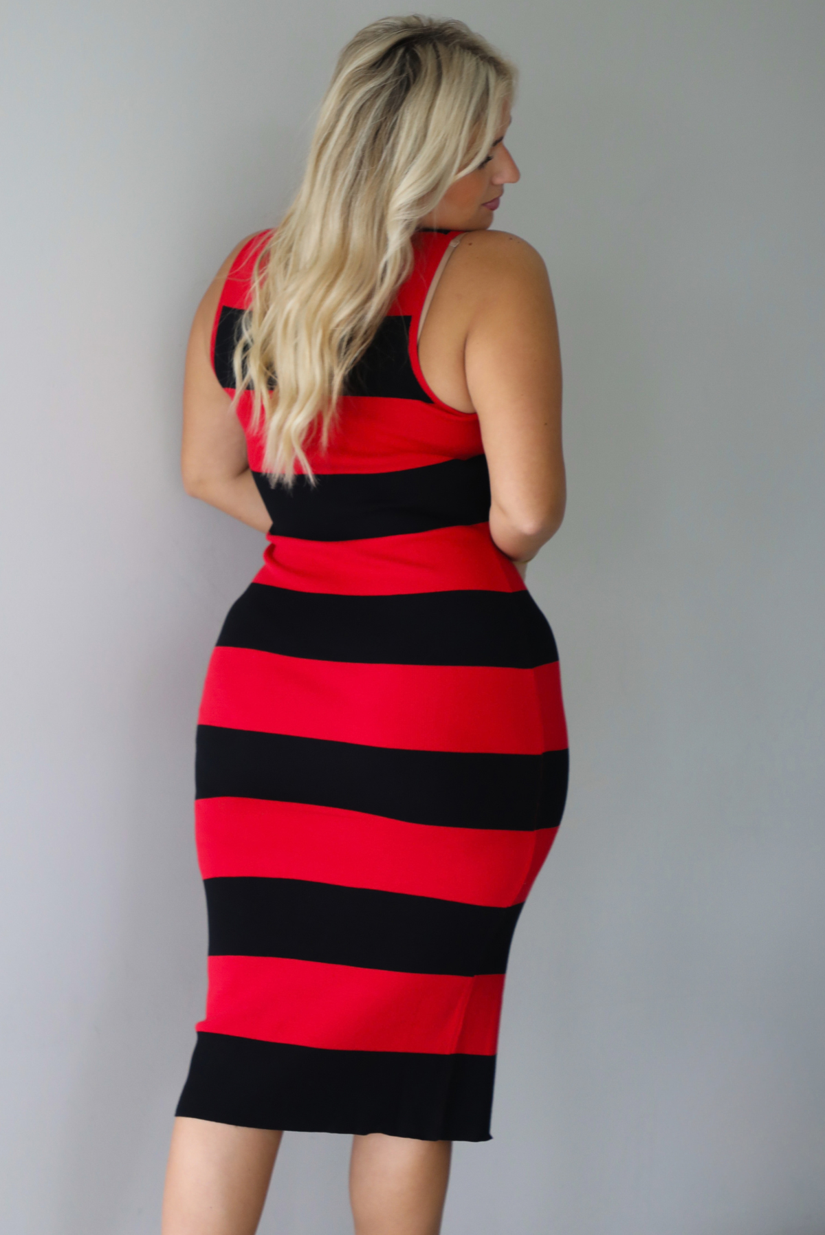 A woman shown from behind wearing a red and black striped body-con midi dress standing indoors against a white wall.
