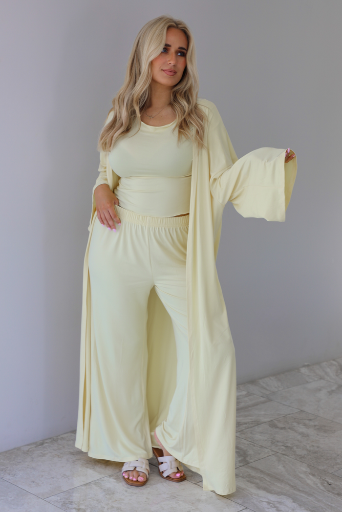 Woman wearing a light yellow three piece set featuring a long sleeved top, long, flared hem pants, and a long, matching cardigan. The photo is taken indoors against a white wall with white marble flooring. 