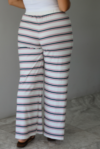 A person shown from the back wearing wide-leg pants with horizontal green and red on a cream-white base, paired with a white top and brown sandals, standing indoors against a white wall with white marble flooring. 