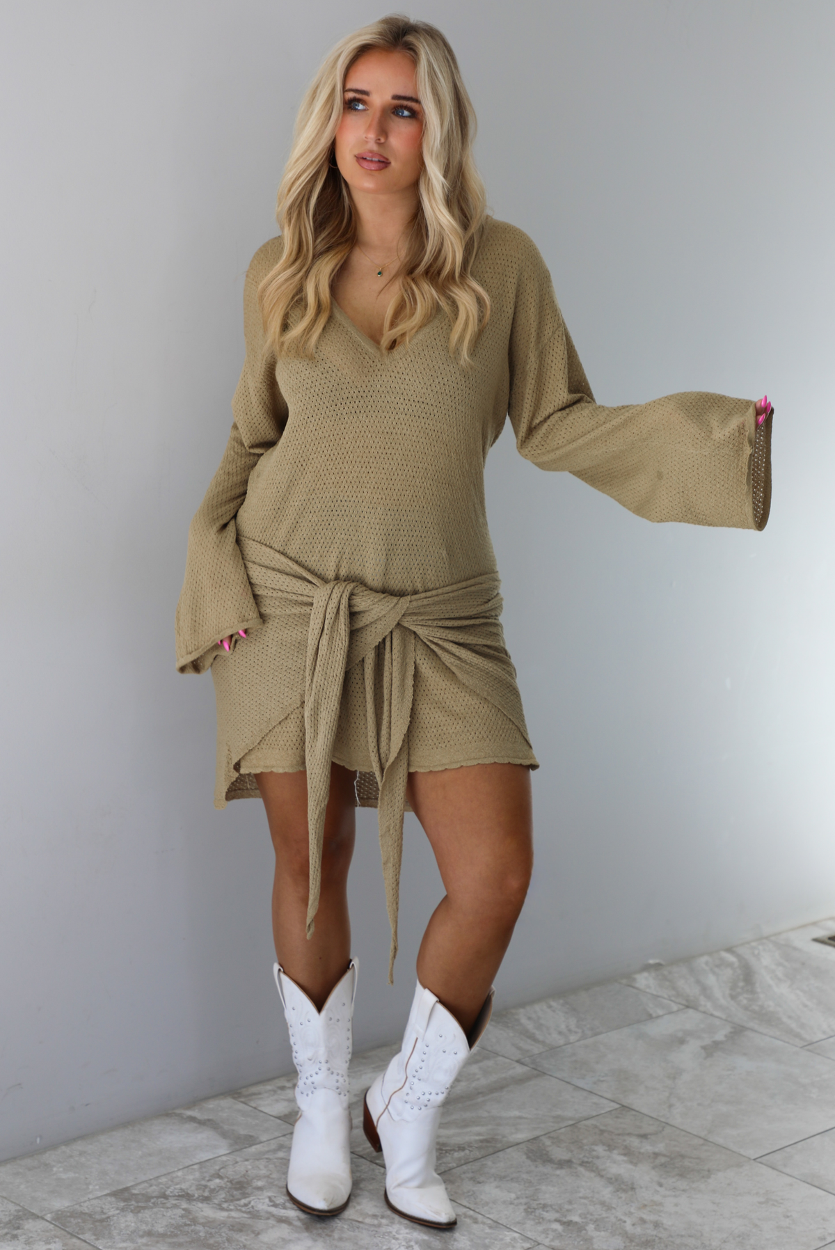 Woman wearing a camel long-sleeve dress that has a tie detail in the front. The dress is paired with white cowboy boots. The photo is taken indoors against a white wall with white marble flooring. 