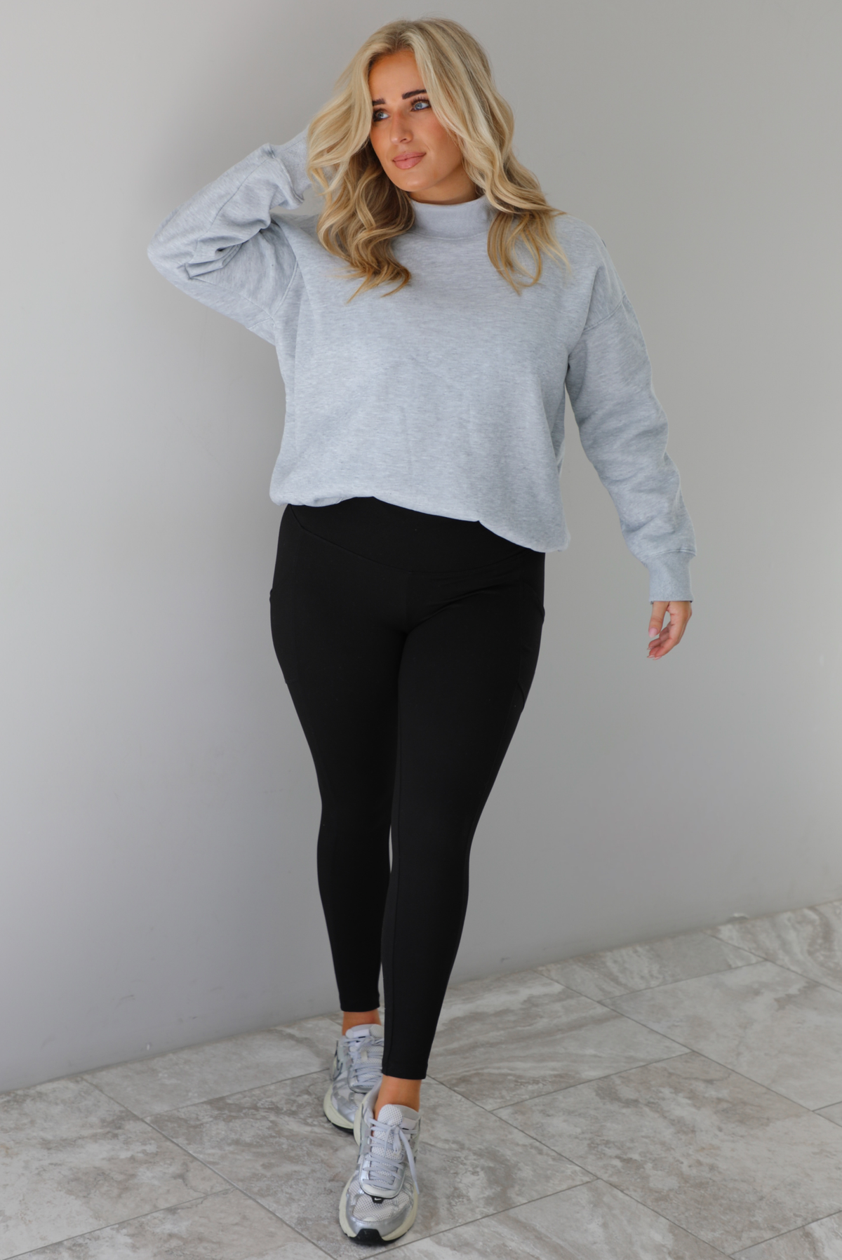 Woman wearing a grey turtleneck sweater tucked into high waisted black leggings. The outfit is paired with silver sneakers. The photo is taken indoors against a white wall with white marble flooring.