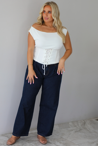 A woman modeling a white off-the-shoulder lace-up corset top paired with dark denim wide-leg jeans and pinkish-brown flat shoes, standing indoors against a white wall with white marble flooring. 