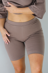 Woman wearing a two piece taupe set featuring an oversized t-shirt and fitted biker shorts.   The photo shows a close up of the biker shorts, which shows a thick band at the top of the waistline, and the fitted design. The photo is taken indoors in front of a white wall.