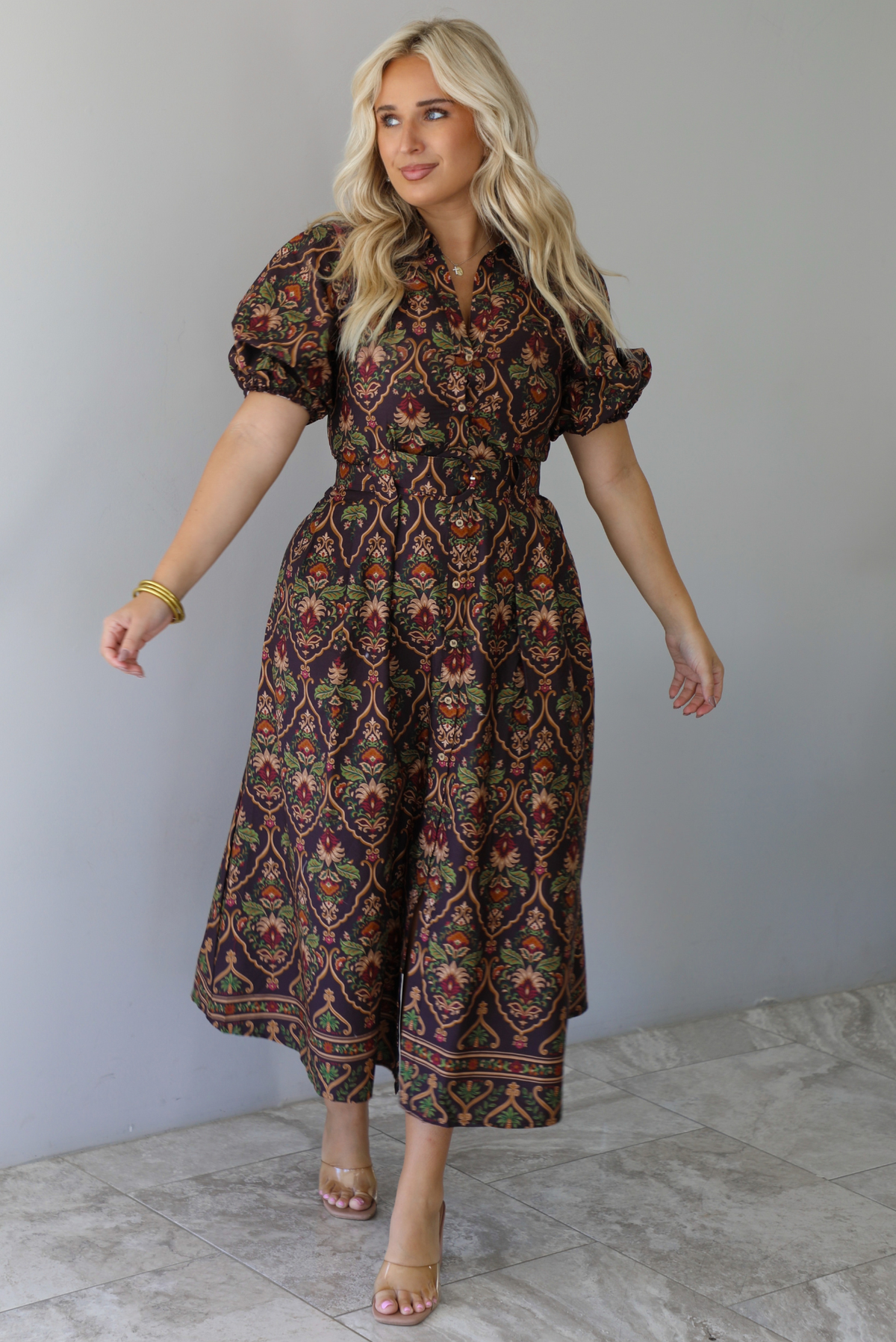 A woman wearing a dark brown midi dress with a multi-colored design throughout, a collared neckline, short puffed sleeves, functional button closures along the front, a belted detail at the waistline, standing indoors against a white wall with white marble flooring. 