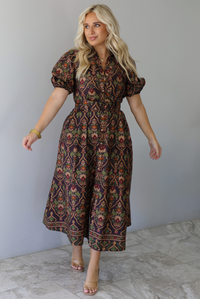 A woman wearing a dark brown midi dress with a multi-colored design throughout, a collared neckline, short puffed sleeves, functional button closures along the front, a belted detail at the waistline, standing indoors against a white wall with white marble flooring. 