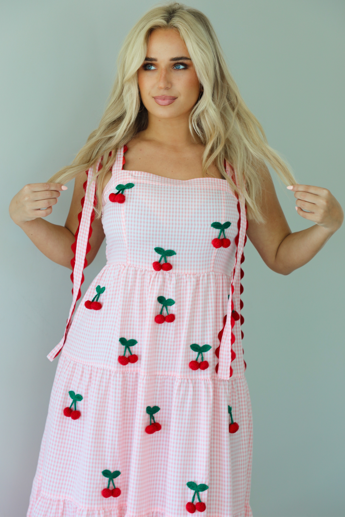 Woman wearing a pink and white gingham  maxi dress with thick straps, a fitted bodice, flowy skirt, and crocheted cherry details on the front indoors against a white wall. The photo shows a closeup of the bodice. 