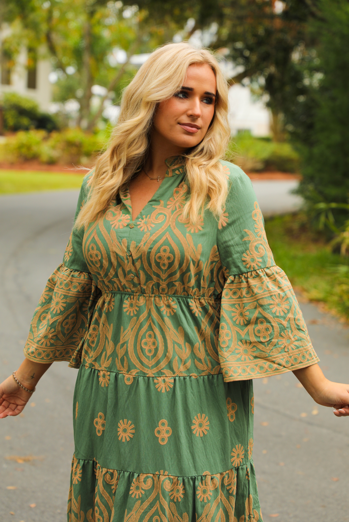 Woman wearing a teal dress that has tan a stitched pattern throughout, quarter length  sleeves, a collared, v-neckline, cinched with elastic waistline, and a maxi-length skirt. The photo shows a close up of the bodice of the dress which has non-functional button closures, and is taken outdoors standing on a paved road with trees and other greenery in the background.
