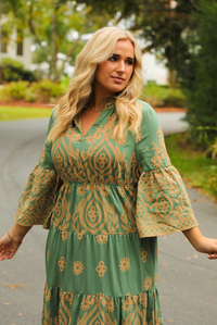 Woman wearing a teal dress that has tan a stitched pattern throughout, quarter length  sleeves, a collared, v-neckline, cinched with elastic waistline, and a maxi-length skirt. The photo shows a close up of the bodice of the dress which has non-functional button closures, and is taken outdoors standing on a paved road with trees and other greenery in the background.