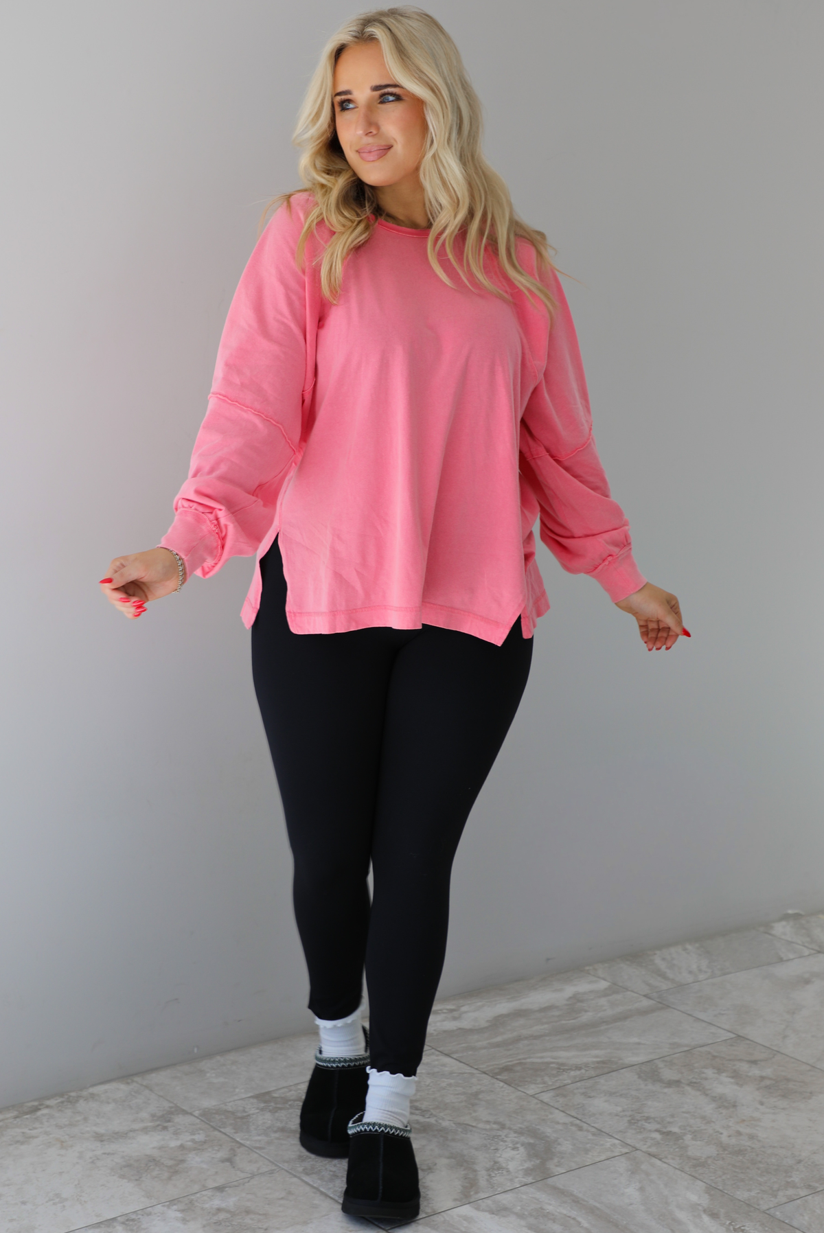 Woman wearing a pink oversized long sleeved top that has a high neckline, exposed seam details, split hem details on either side, and is lightweight. The top is modeled with a pair of fitted black leggings with black uggs and socks. The photo is modeled indoors against a white wall with white marbled flooring. 