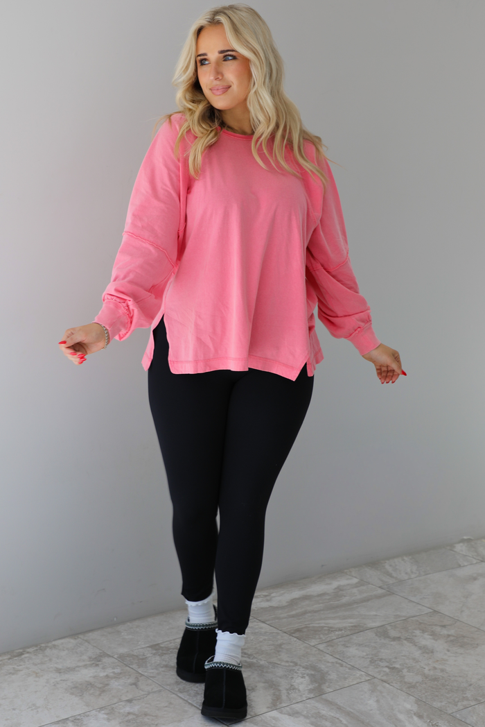 Woman wearing a pink oversized long sleeved top that has a high neckline, exposed seam details, split hem details on either side, and is lightweight. The top is modeled with a pair of fitted black leggings with black uggs and socks. The photo is modeled indoors against a white wall with white marbled flooring. 