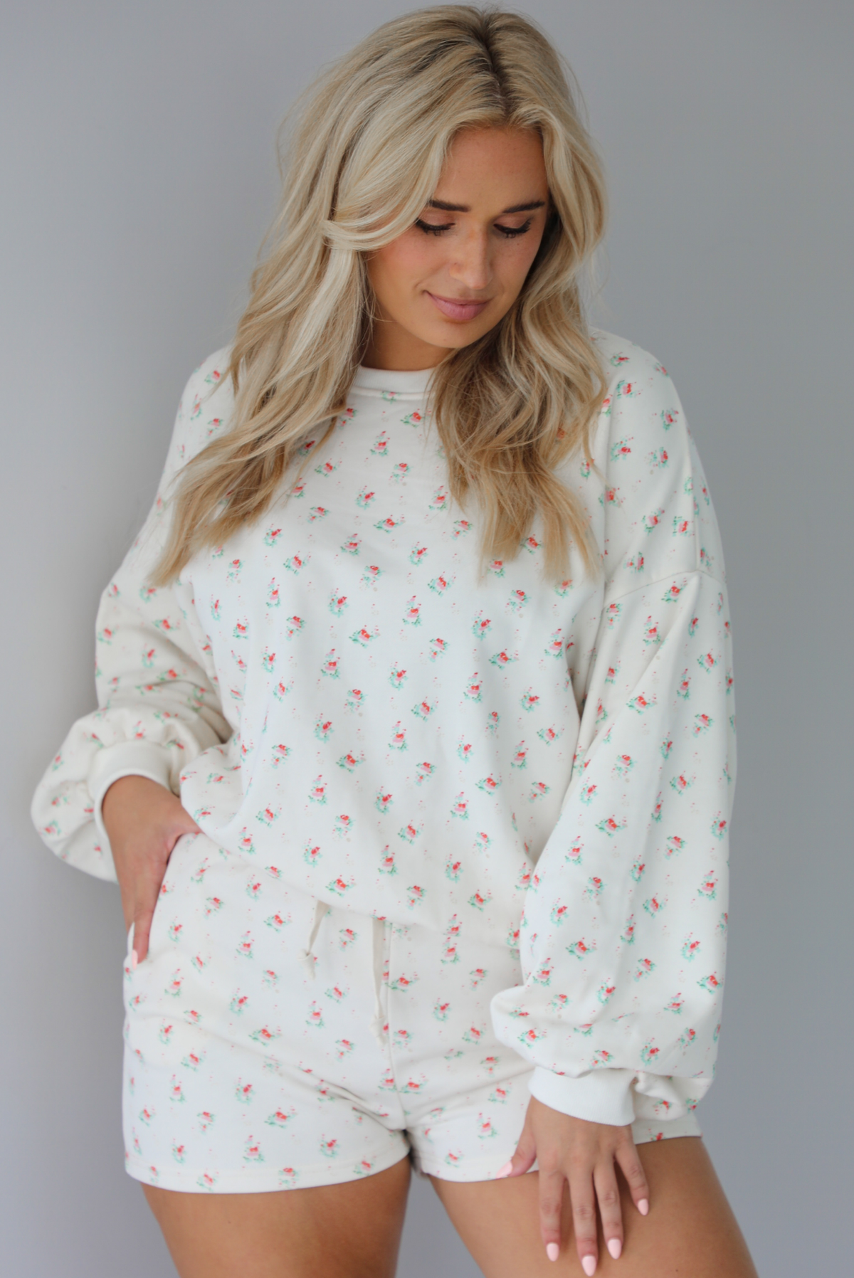 Woman wearing a two piece cream set that has a floral design throughout featuring a sweatshirt and sweat-shorts indoors against a white wall.