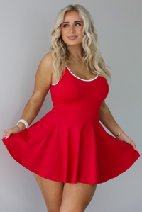 Woman wearing a red athleisure dress that has white trim at the top of the bodice, a fitted bodice, and flowy skirt. The photo is taken indoors against a white wall.
