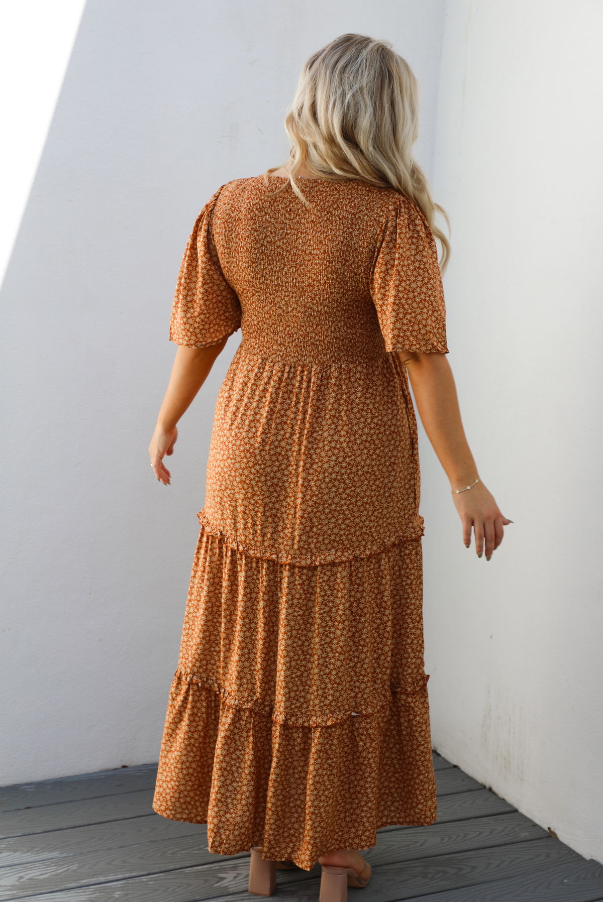 Back of a woman model outdoors wearing a rust dress with a cream floral print pattern throughout. It has flowy short sleeves, a smocked bodice, and a flowy maxi length skirt with functional pockets on either side. 