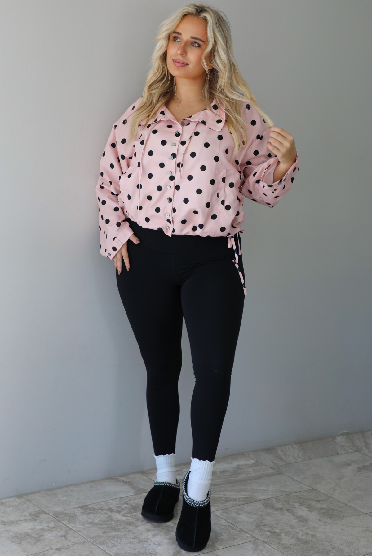 A woman wearing a pink button-down jacket with a black polka dot print pattern, paired with black leggings, white ankle socks, and black slide on slippers, standing indoors against a white wall with white marble flooring. 