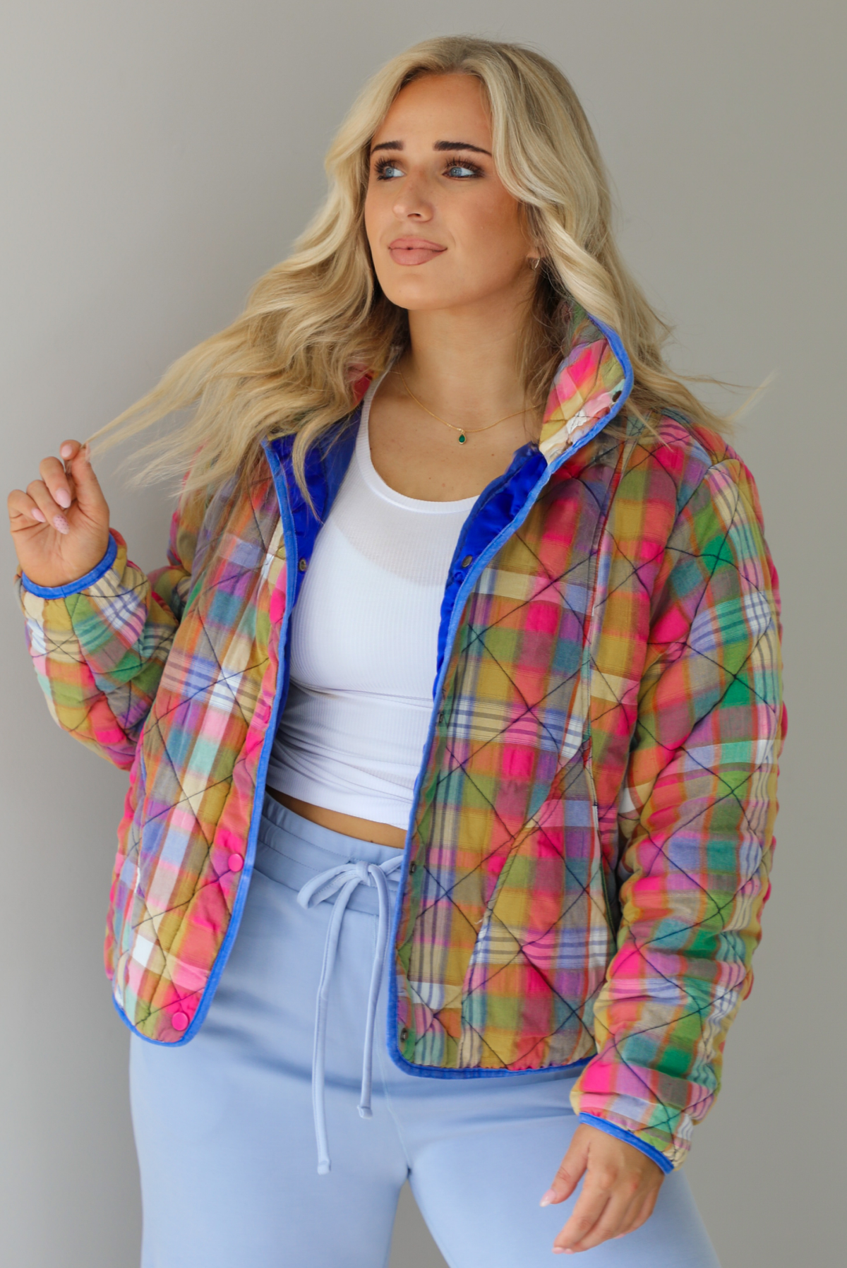 Woman wearing a multi-colored plaid, quilted style jacket paired with a white top underneath and high waisted blue lounge pants. The photo shows a closeup of the jacket, with the functional button closures in the front, functional pockets, and quilted texture. The photo is taken indoors against a white wall.