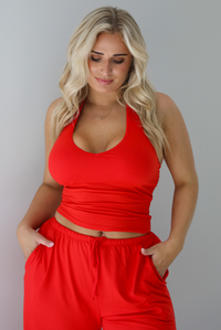 Woman wearing a red two piece set featuring a  tank top and slouchy pants. The photo shows a closeup of the tank top and the upper portion of the pants. The photo is taken indoors against a white wall.