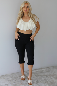 Woman wearing a cream ruffled cropped top with high waisted black, textured capri pants indoors against a white wall with white marble flooring. 