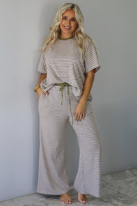 A woman modeling a matching two-piece loungewear set consisting of a short-sleeve top and wide-leg pants, both featuring thin beige and olive green horizontal stripes, with an olive green drawstring waist, standing indoors against a white wall with white marble flooring. 
