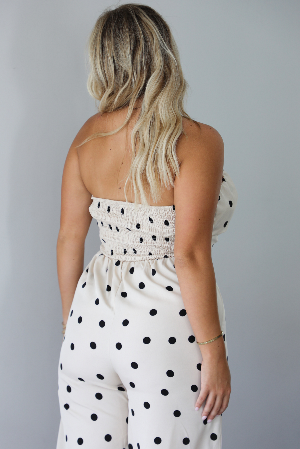 A woman shown from behind wearing a sand strapless jumpsuit with a black polka dot print pattern throughout, standing indoors against a white wall. The photo focuses on the upper half of the jumpsuit, which has a smocked design. 
