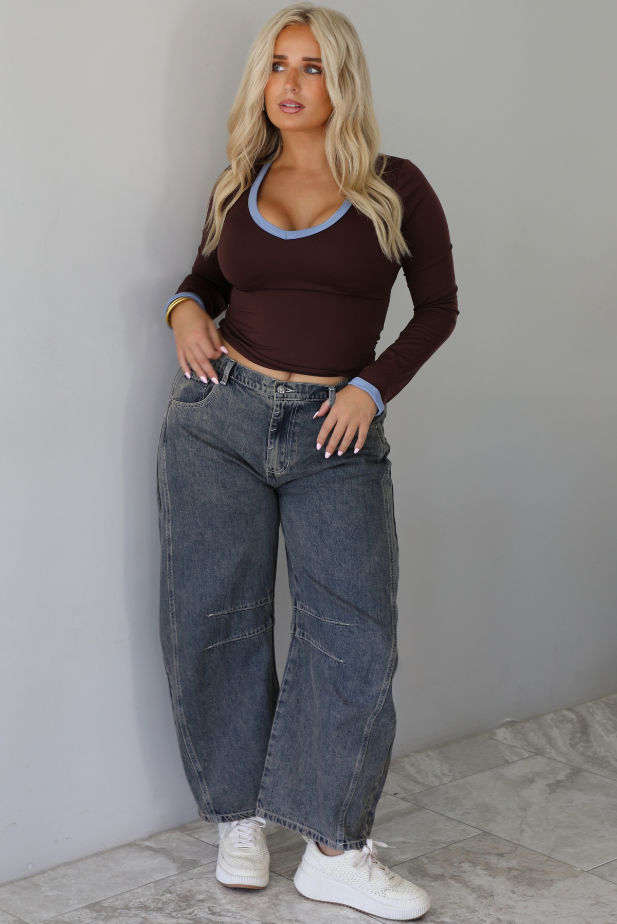 A woman wearing a dark brown, fitted, long sleeved, slightly cropped top with blue trim detailing at the v-neckline and at the sleeve hems, paired with dark acid washed barrel denim jeans, standing indoors in front of a white wall with white marble flooring.