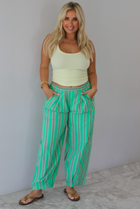 A woman wearing a pale yellow tank and mid-rise pants that have a green, brown, lime green, and tan striped pattern, which have an elastic waistline, functional pockets on either side, and cinched details at the ankles, standing indoors against a white wall with white marble flooring. 