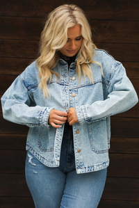 Woman model wearing an oversized, acid washed, light denim jacket, paired with a sheer mesh leopard print top, and medium wash denim jeans. The photo shows the model holding the two sides of the jacket together to show how it would look buttoned up.