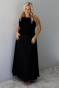 A woman wearing a two piece black set featuring a smocked detail top and high waisted maxi skirt, standing indoors against a white wall with white marble flooring. 