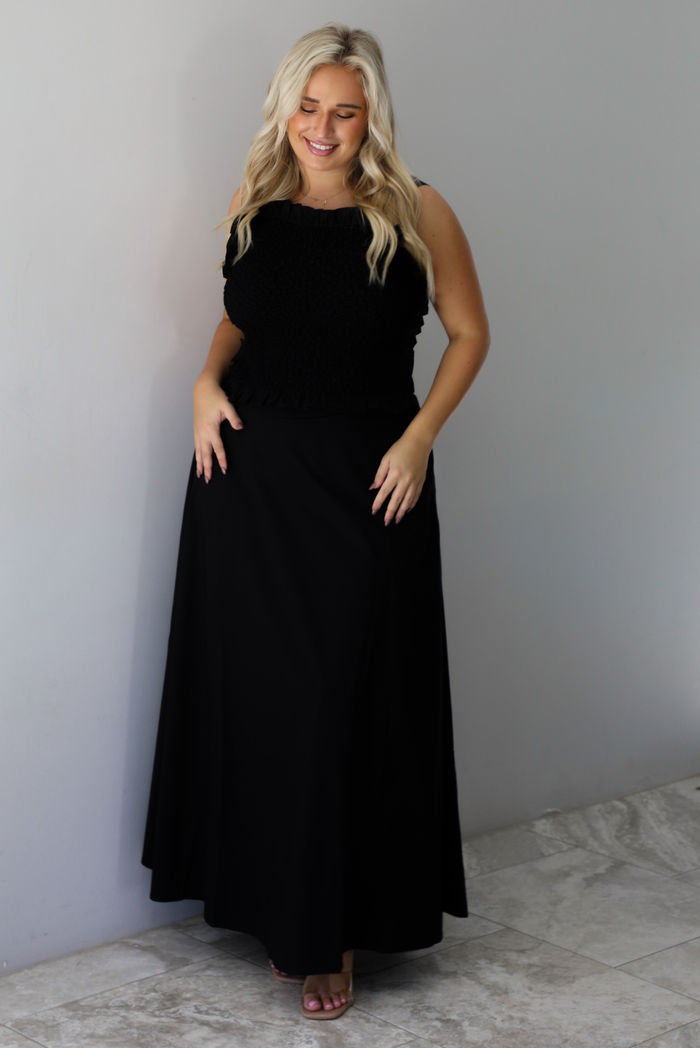 A woman wearing a two piece black set featuring a smocked detail top and high waisted maxi skirt, standing indoors against a white wall with white marble flooring. 
