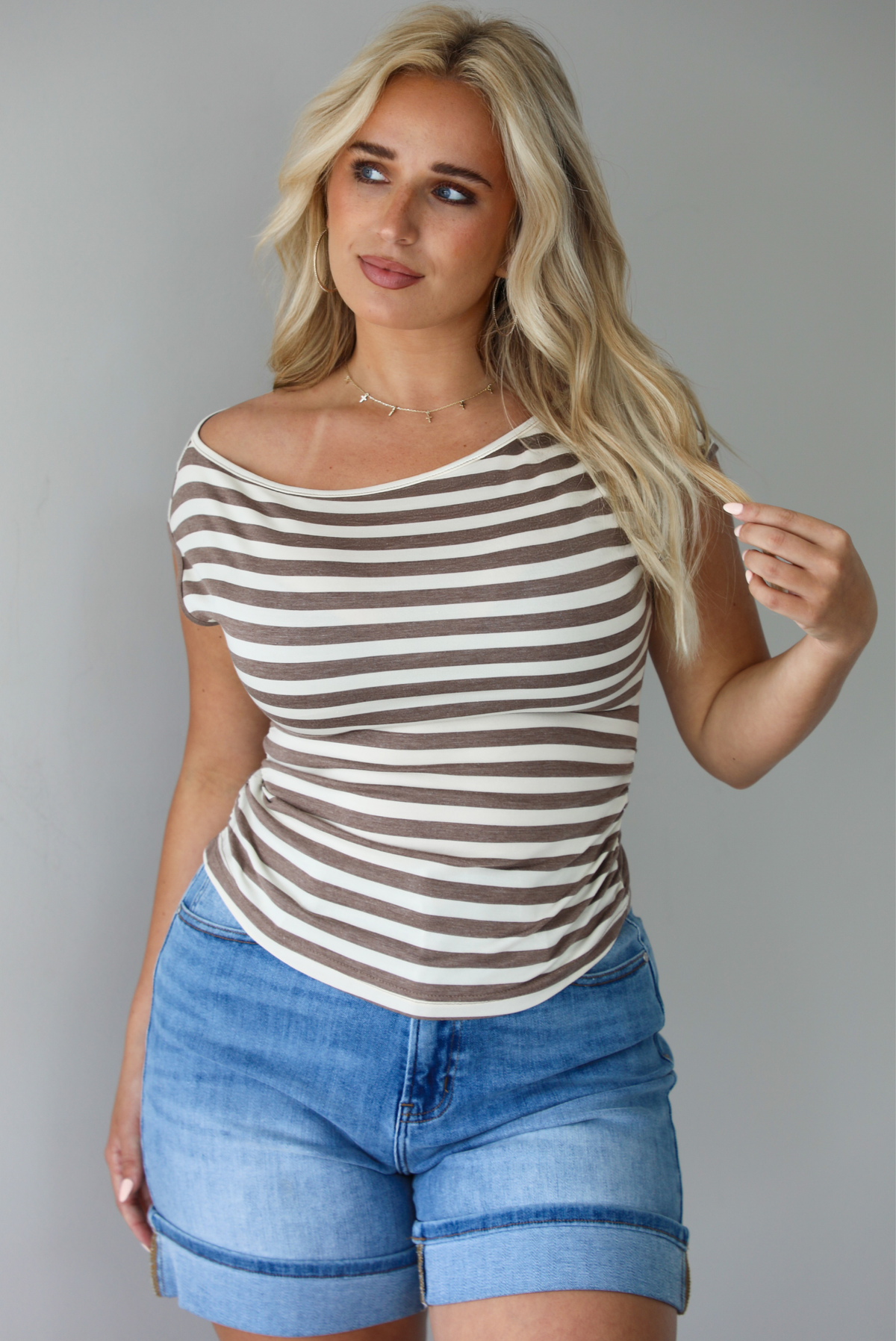 Woman wearing a mocha and white striped top with an off the shoulder design with medium wash denim shorts indoors against a white wall.