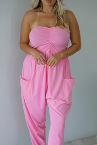 Woman wearing a pink strapless jumpsuit with a fitted bodice and slouchy pants indoors against a white wall. The photo shows a closeup of the bodice and the functional pockets at the hips. 