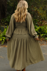 The back of a woman model wearing an olive colored maxi length dress that has a smocked bodice, long sleeves that cinch with elastic at the wrists, and a flowy skirt. The photo is taken outdoors with greenery in the back.