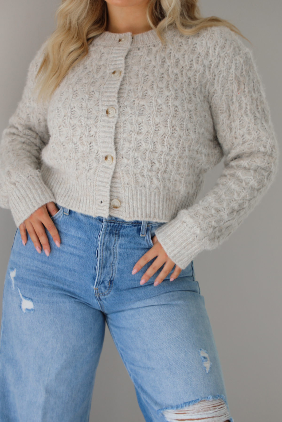 Woman wearing a light grey cardigan buttoned up with high waisted flared hem medium wash denim. The photo shows a closeup of the texture of the cardigan, which resembles a loose textured cable knit. The photo is taken indoors against a white wall.