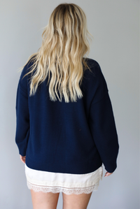 A woman shown from behind wearing a navy sweater dress with a cream lace trim detail peaking out from underneath the dress giving it a layered look, standing indoors against a white wall.