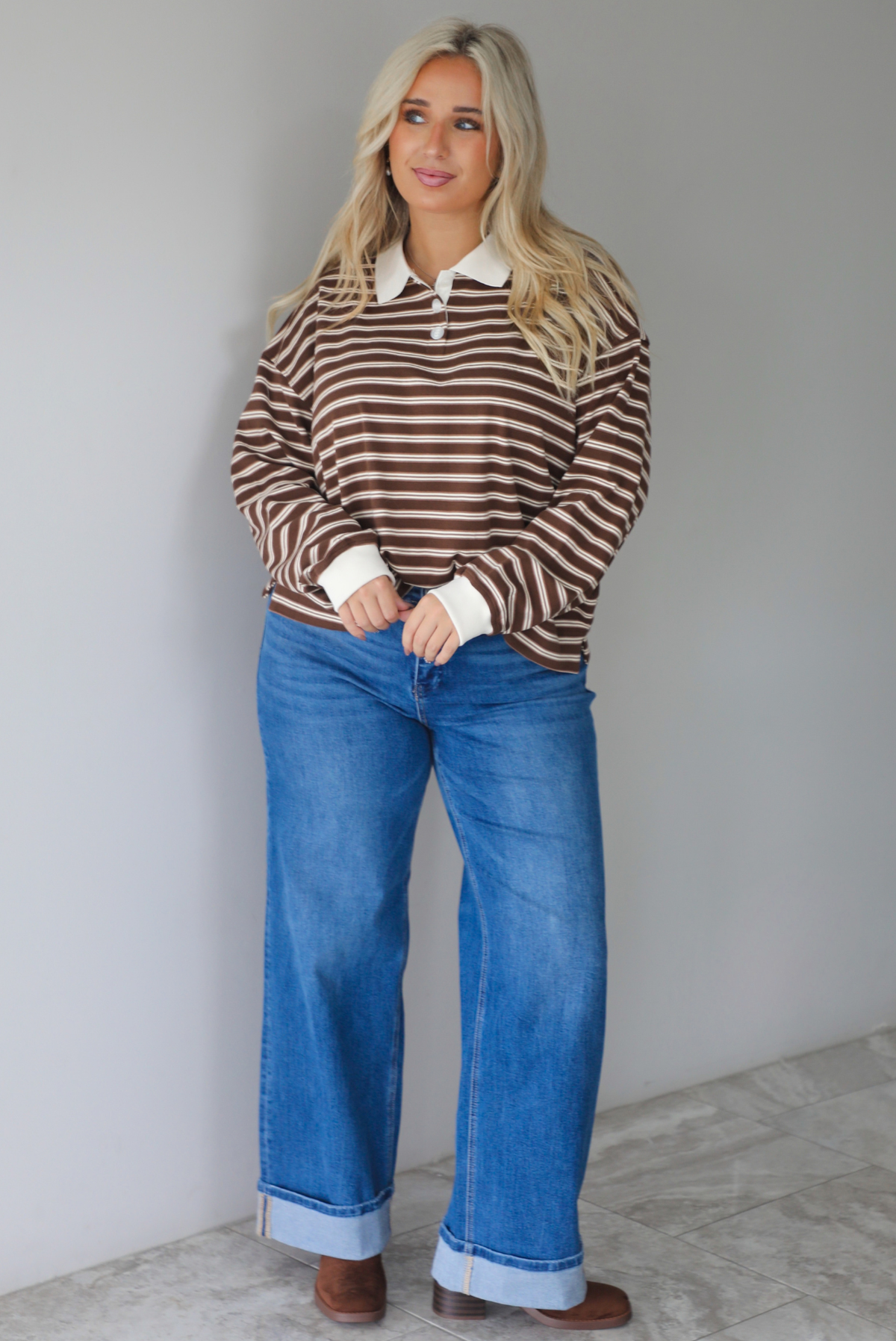 A woman wearing a brown and cream striped collared quarter-button top, paired with dark, wide leg denim with a cuffed hem, standing indoors against a white wall with white marble flooring. 