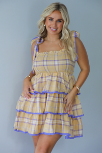 A woman modeling a yellow and purple plaid tiered mini dress with functional tie straps, standing indoors against a white wall.