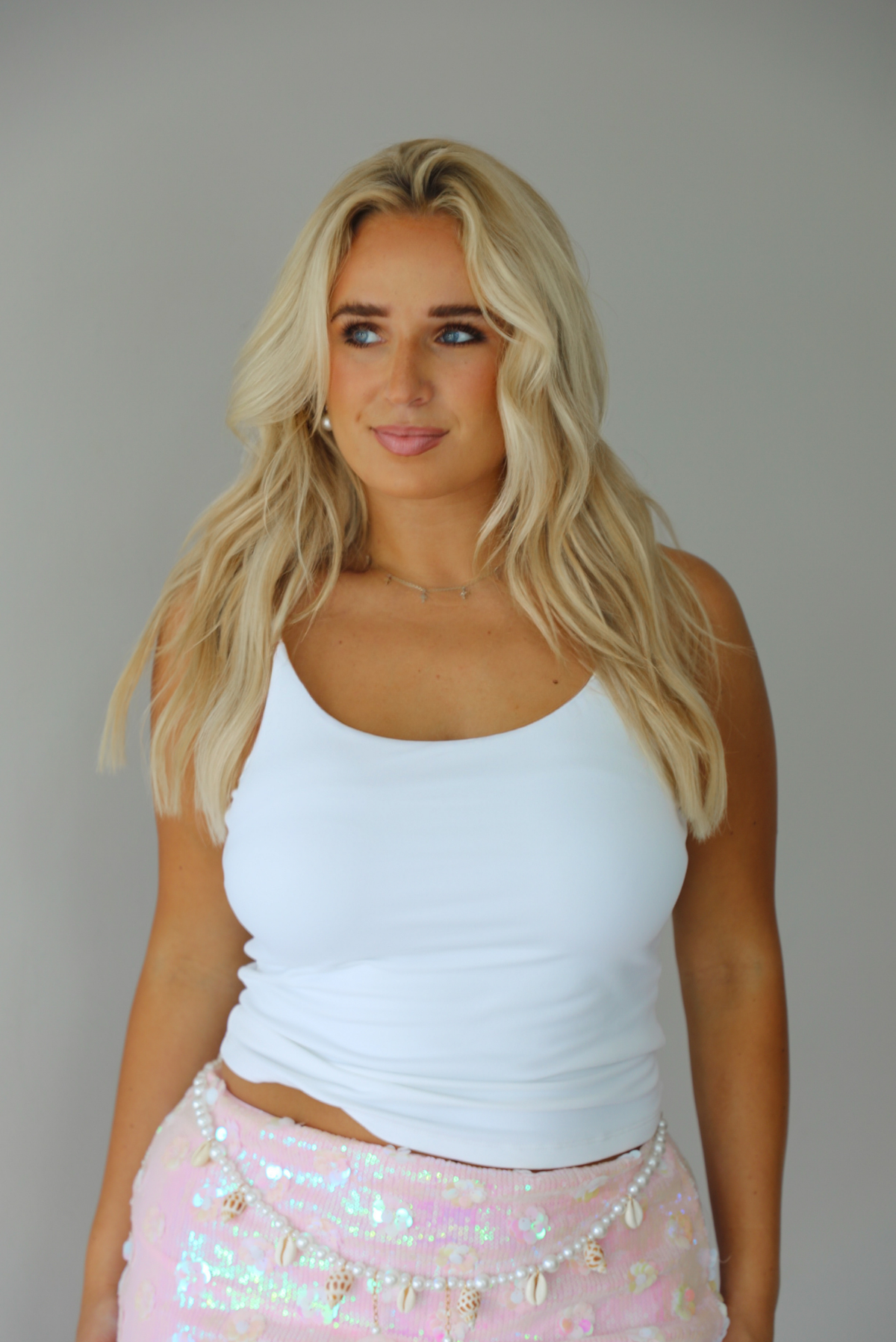 A blonde woman wearing a white ribbed tank top and a light pink sequined skirt with gold fringe, posing against a solid gray studio background.