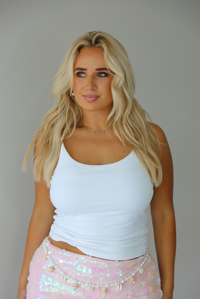 A blonde woman wearing a white ribbed tank top and a light pink sequined skirt with gold fringe, posing against a solid gray studio background.