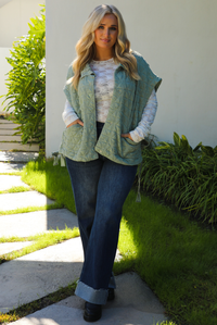 Woman model wearing a sage colored oversized vest paired with a white lace long sleeved top and dark denim outdoors.  The vest has functional pockets on either side, functional button closures in the front, and an oversized fit. 