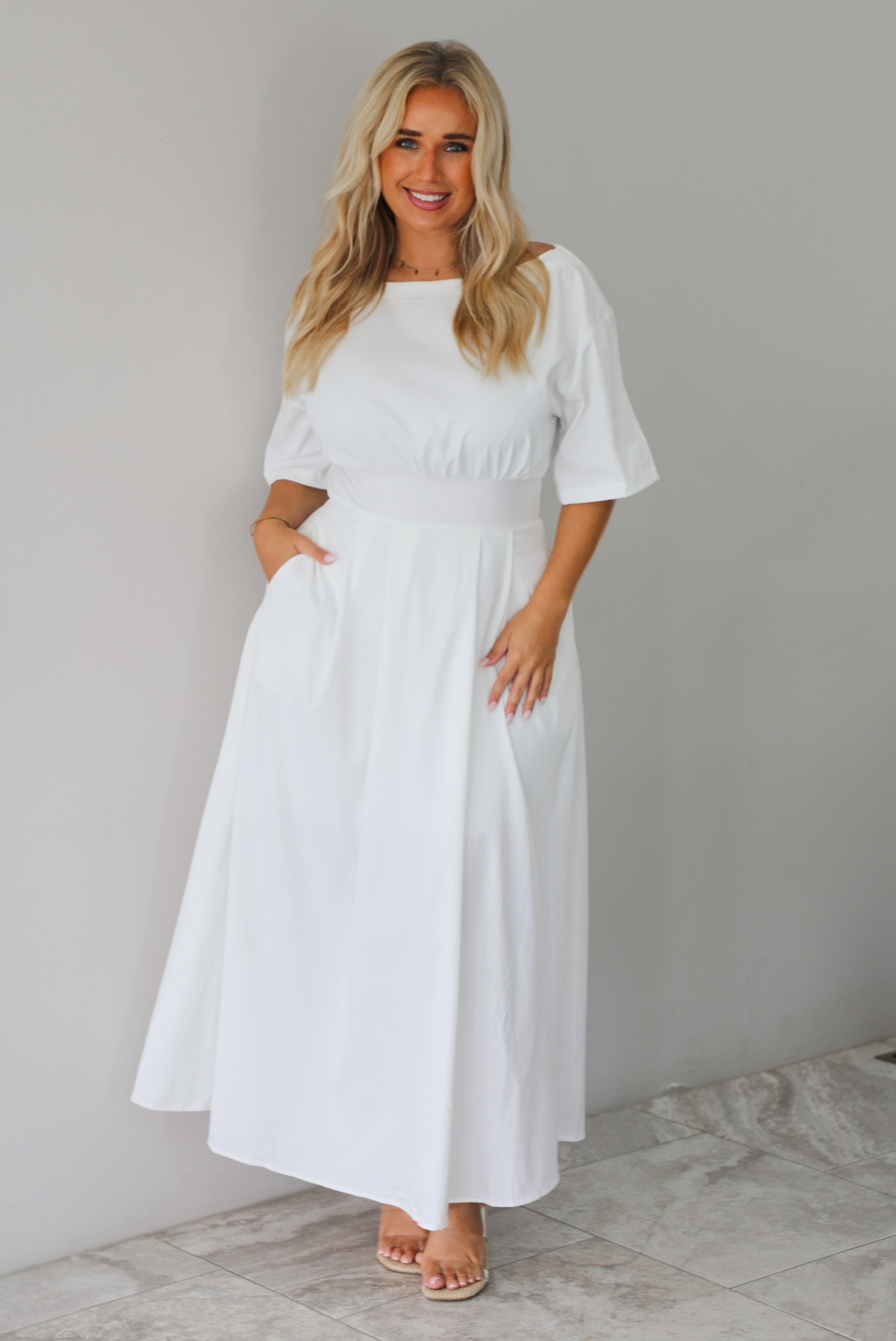 A woman modeling a white midi dress with short puff sleeves, a wide boat neckline, and side pockets, standing indoors against a white wall with white marble flooring. 