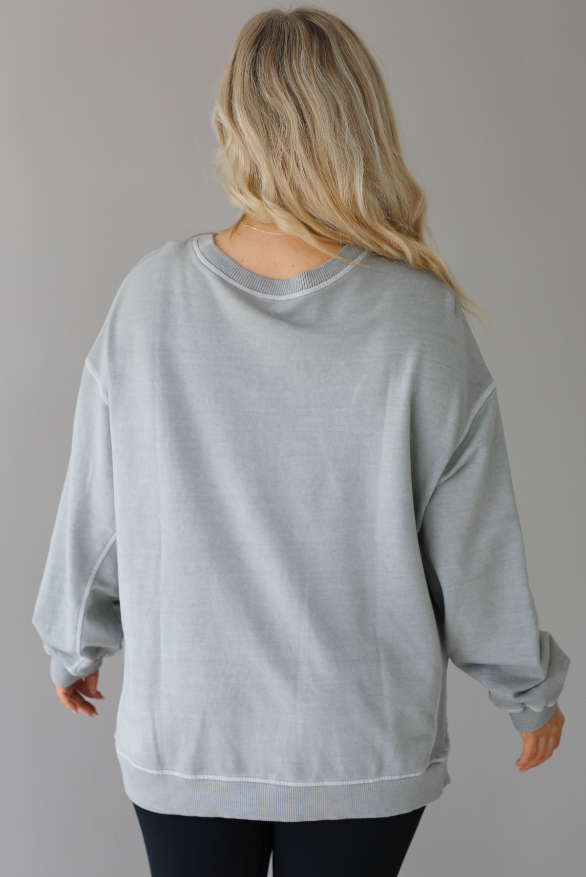 Woman wearing a grey oversized sweater with black leggings that have a slightly flared hem with split hem details in the front. The photo shows a closeup of the back of the sweater and has a white background. 